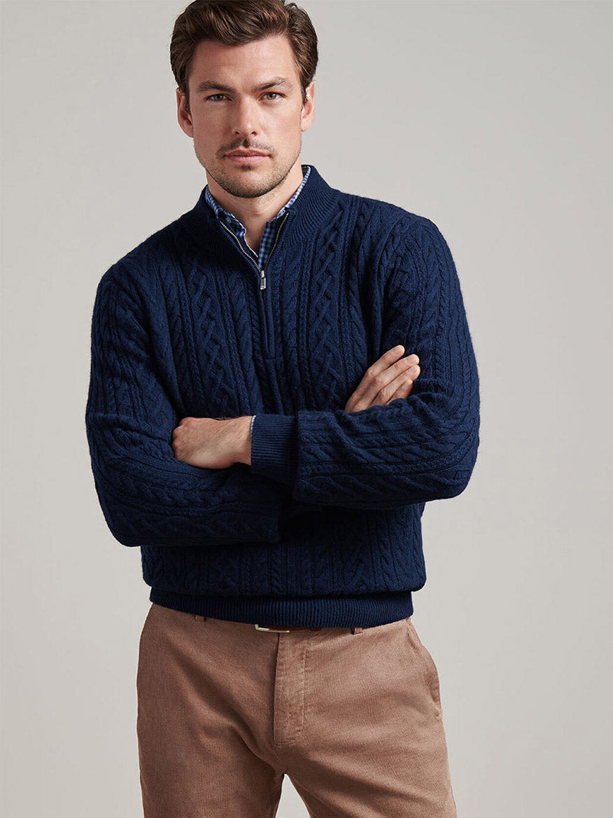 Man wearing the Peter Millar Crescent Ridge Cable Quarter-Zip Sweater in Navy over a blue collared shirt and tan pants, standing with arms crossed against a plain background.
