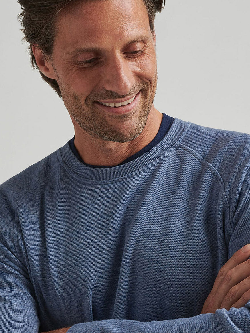 A man with brown hair and stubble smiles while looking down, arms crossed, wearing the Peter Millar Crown Comfort Knit Crew in Blue Freesia over a dark shirt.