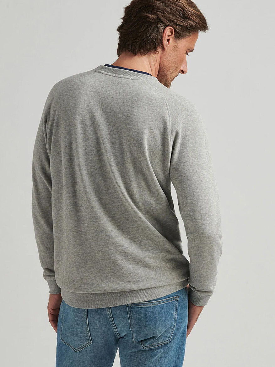 A man wearing the Peter Millar Crown Comfort Knit Crew in Light Grey and blue jeans stands with his back to the camera against a plain light background.
