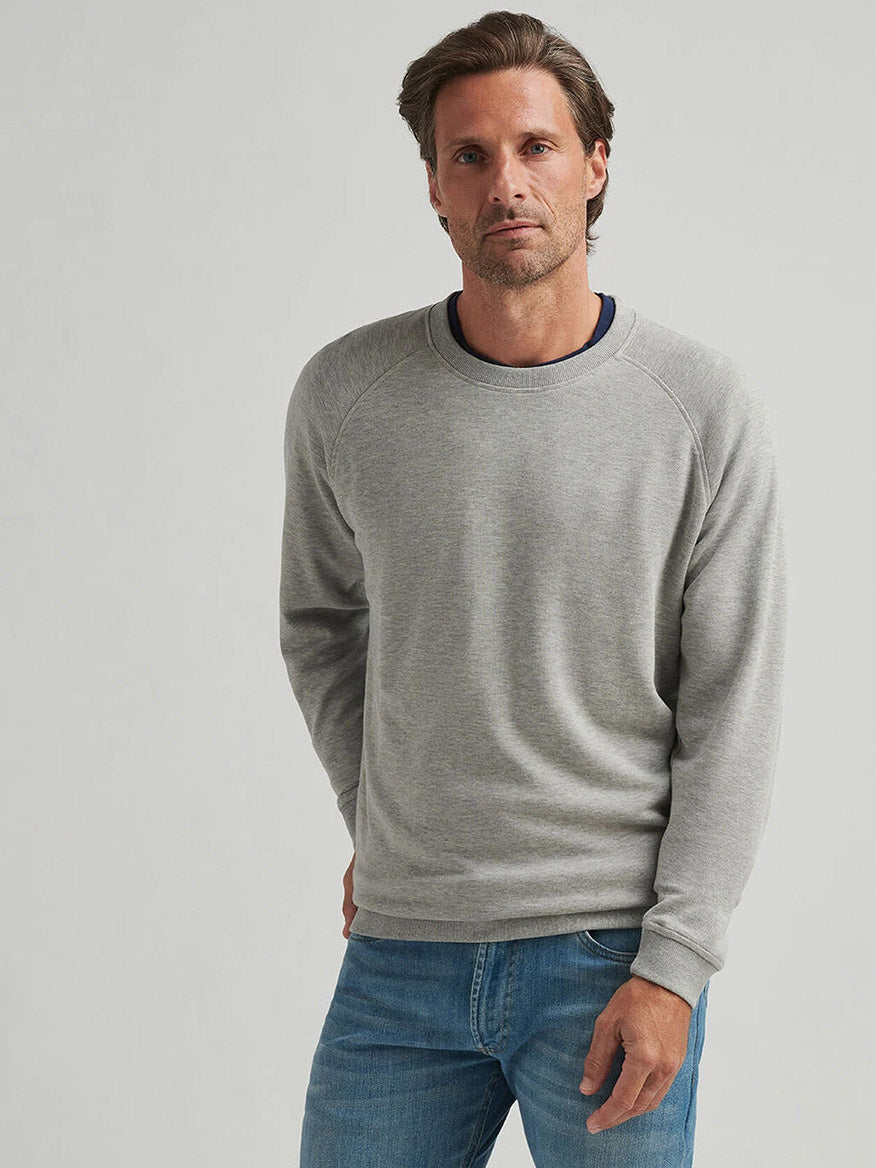 A man with light brown hair wears the Peter Millar Crown Comfort Knit Crew in Light Grey and blue jeans, standing against a plain background to showcase timeless style.