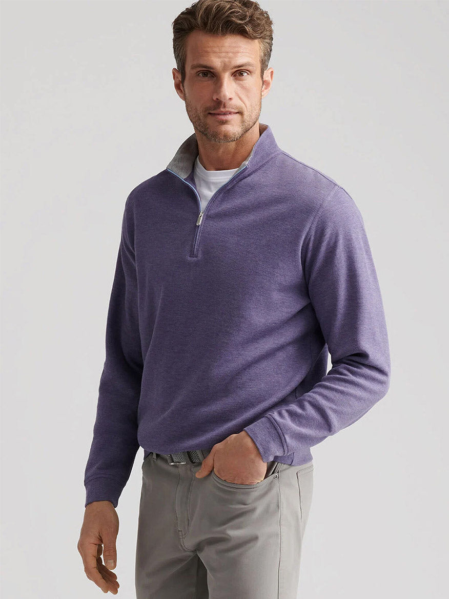 A man stands against a plain light background wearing the Peter Millar Crown Comfort Pullover in Deep Violet over a white t-shirt and gray pants.