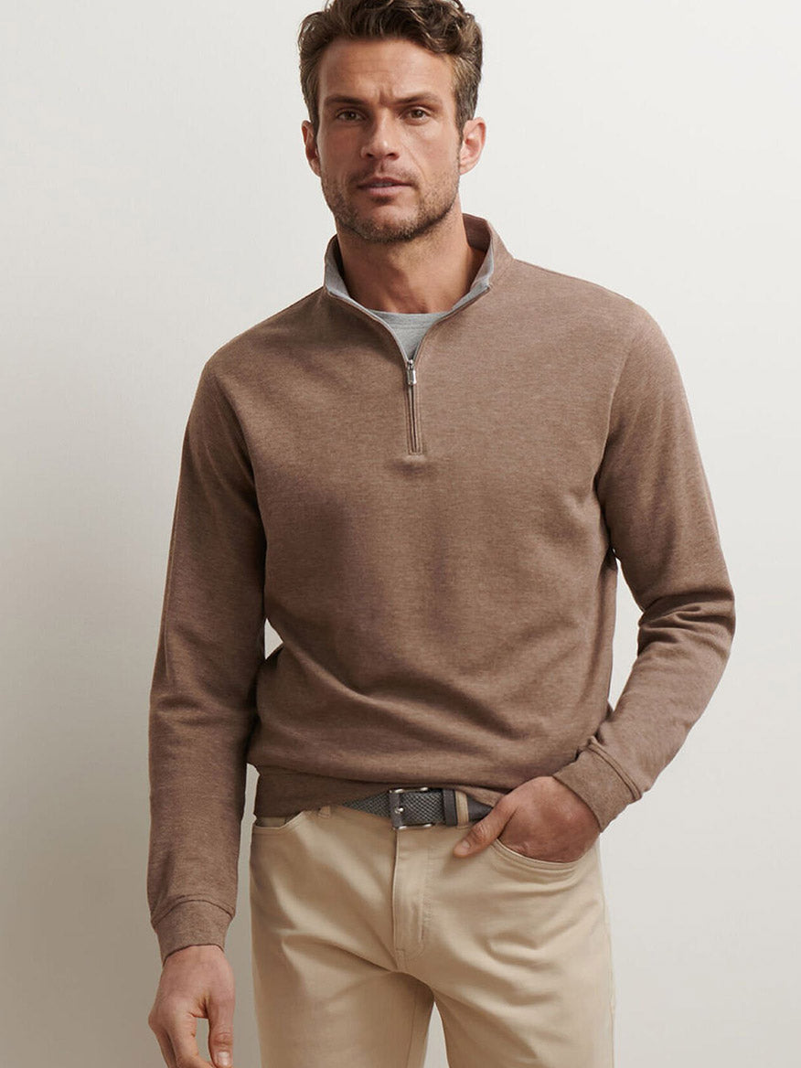A man stands against a plain background wearing the Peter Millar Crown Comfort Pullover in Poplar Brown and beige pants, with one hand in his pocket.