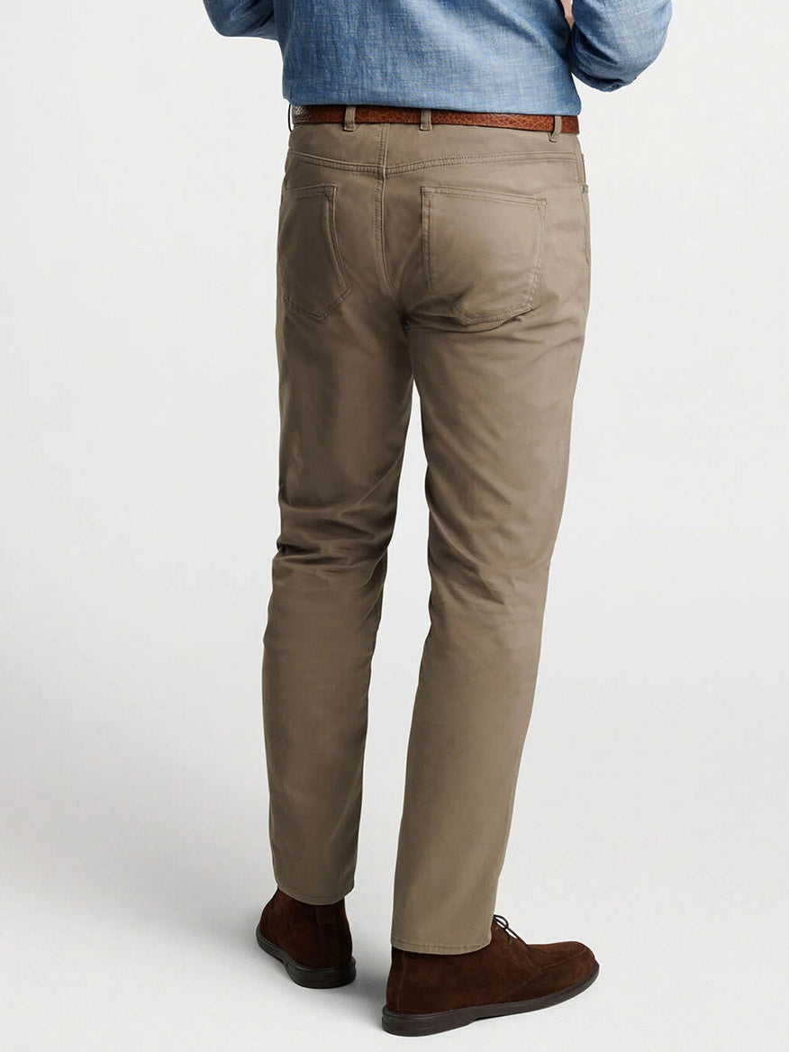 Man stands with his back to the camera, wearing Peter Millar Delvin Twill Five-Pocket Pant in Rye, a blue shirt, brown belt, and brown shoes against a plain background.