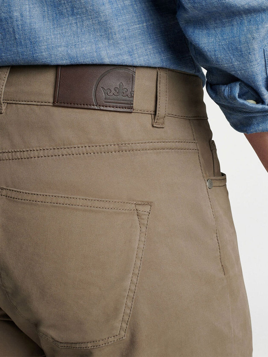 Close-up of a person wearing the Peter Millar Delvin Twill Five-Pocket Pant in Rye, styled with a blue shirt and brown leather belt, highlighting the back pocket and branded patch.