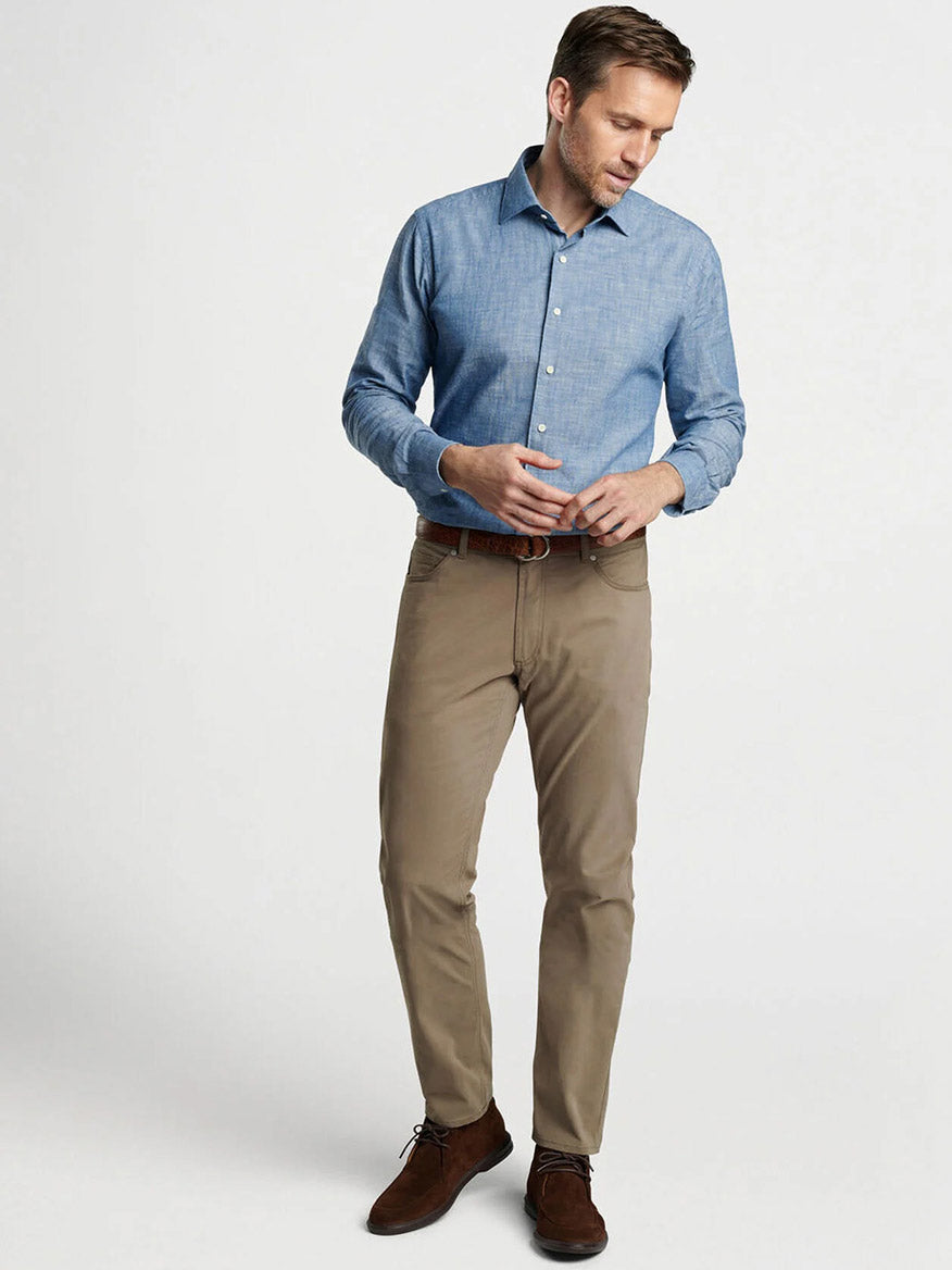 A man stands against a plain, light background wearing tan Peter Millar Delvin Twill Five-Pocket Pant in Rye, a light blue brushed twill button-up shirt, and brown shoes.