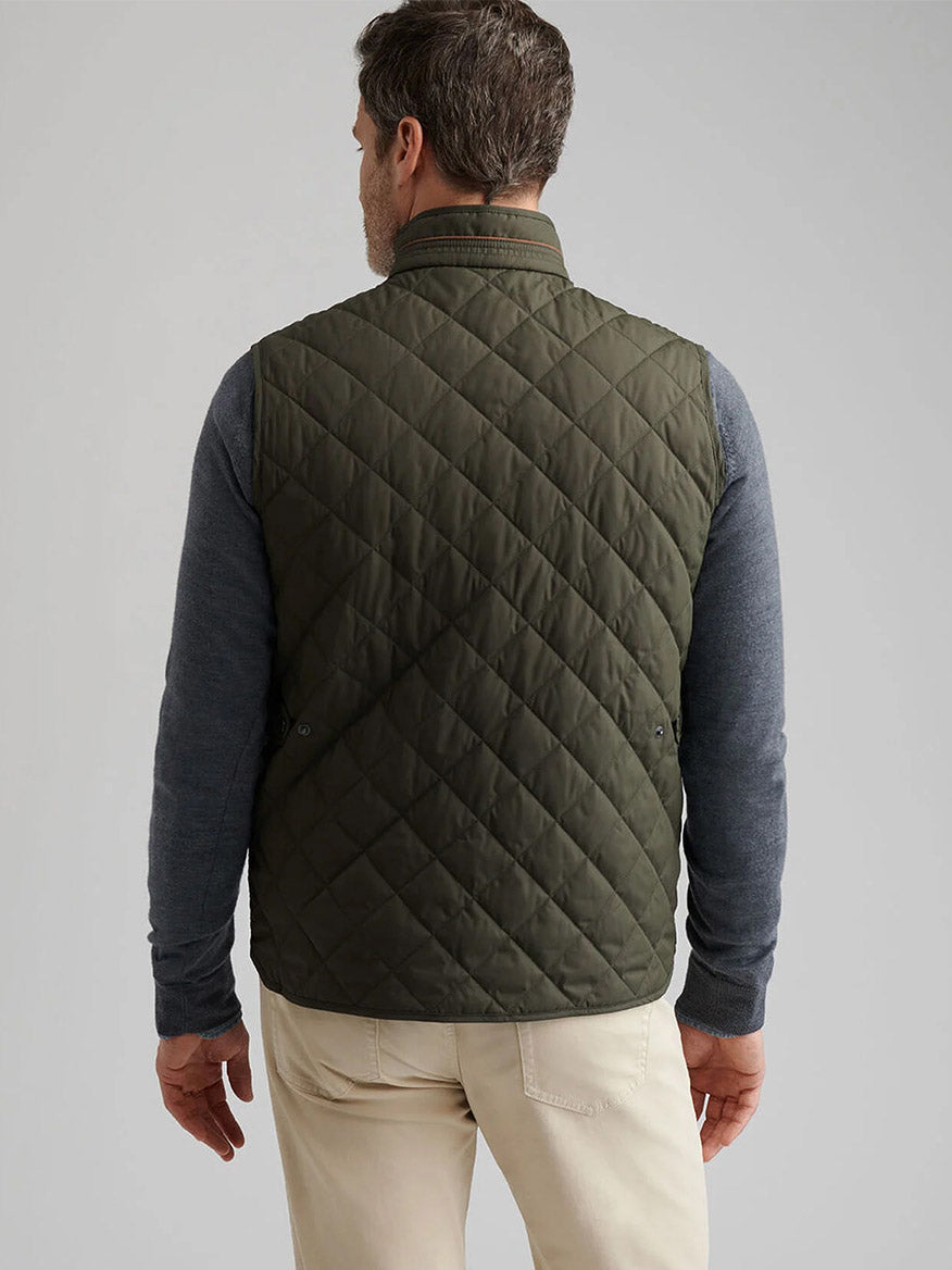 A man is seen from behind wearing the Peter Millar Essex Vest in Dark Olive over a gray long-sleeve shirt and beige pants against a plain background.