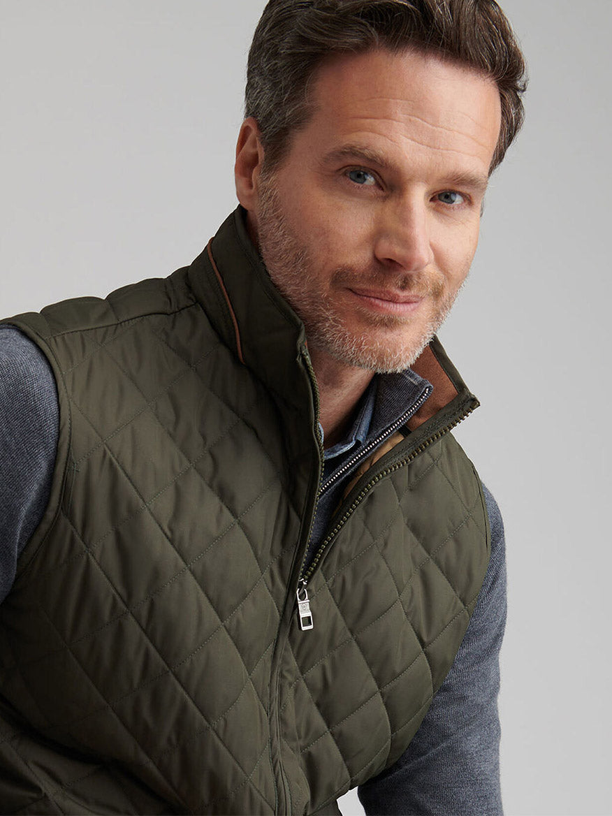 A man with short brown hair and light stubble wears the Peter Millar Essex Vest in Dark Olive over a gray zip-up sweater, looking at the camera against a plain background.