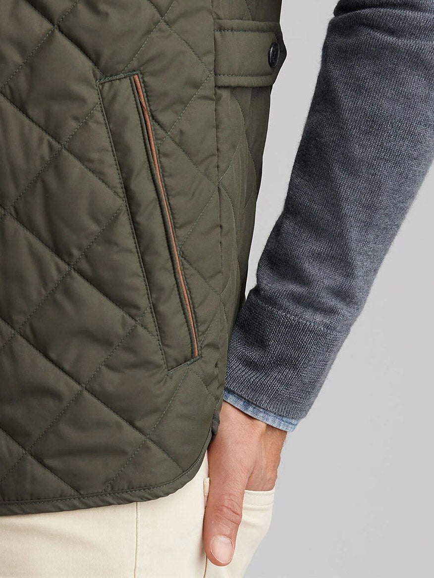 A person wearing the Peter Millar Essex Vest in Dark Olive, a blue-gray sweater, and beige pants with their hand resting by their side.