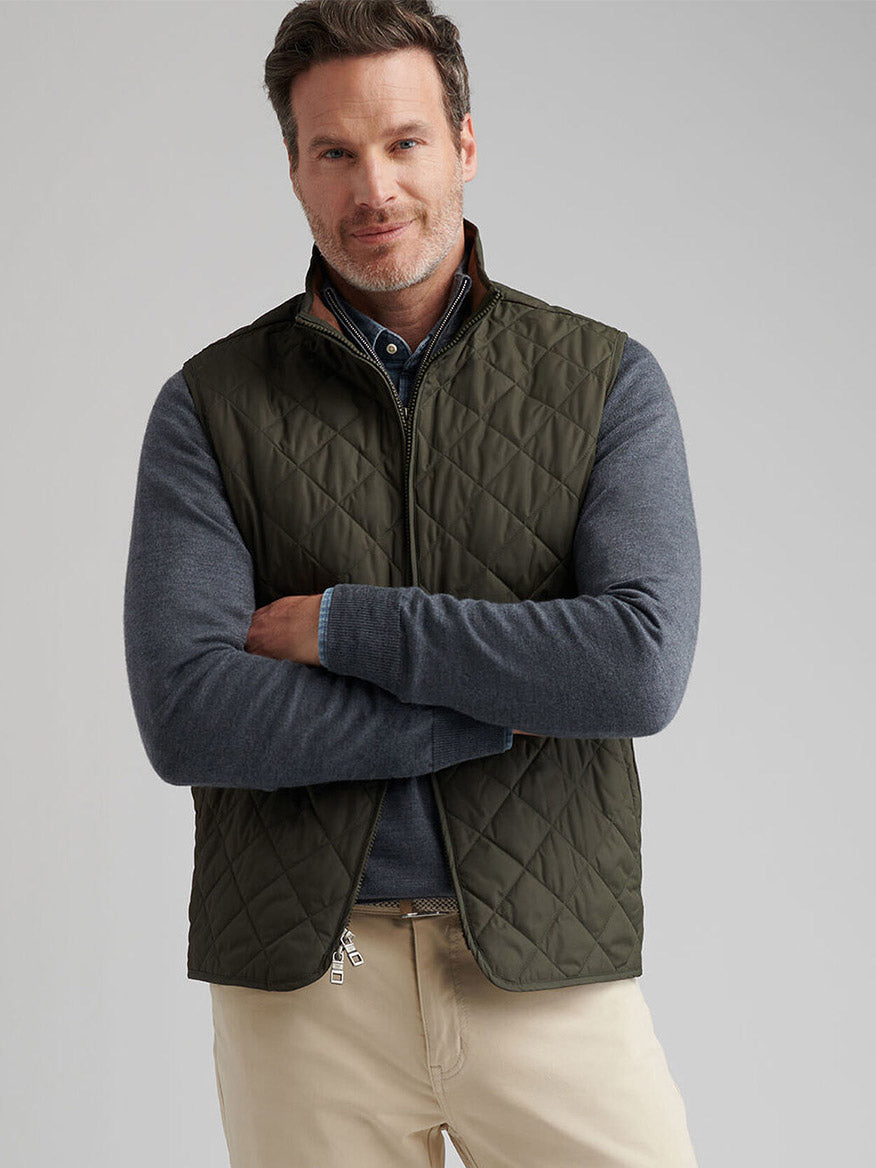 A man with short hair and a trimmed beard stands with arms crossed against a plain background, wearing the Peter Millar Essex Vest in Dark Olive over a gray long-sleeve shirt and beige pants.