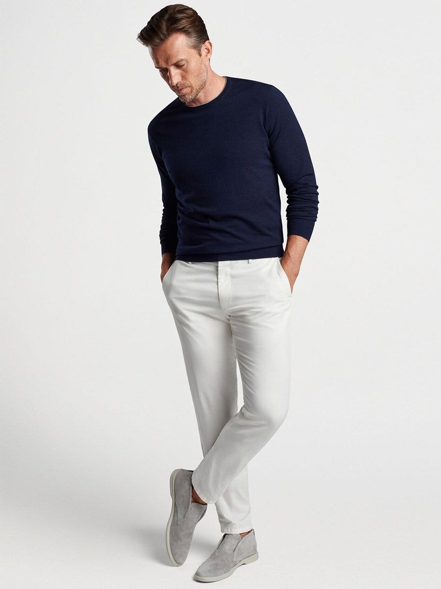 A man in a Peter Millar Excursionist Flex Crew in Navy sweater and white pants posing with one hand in his pocket.