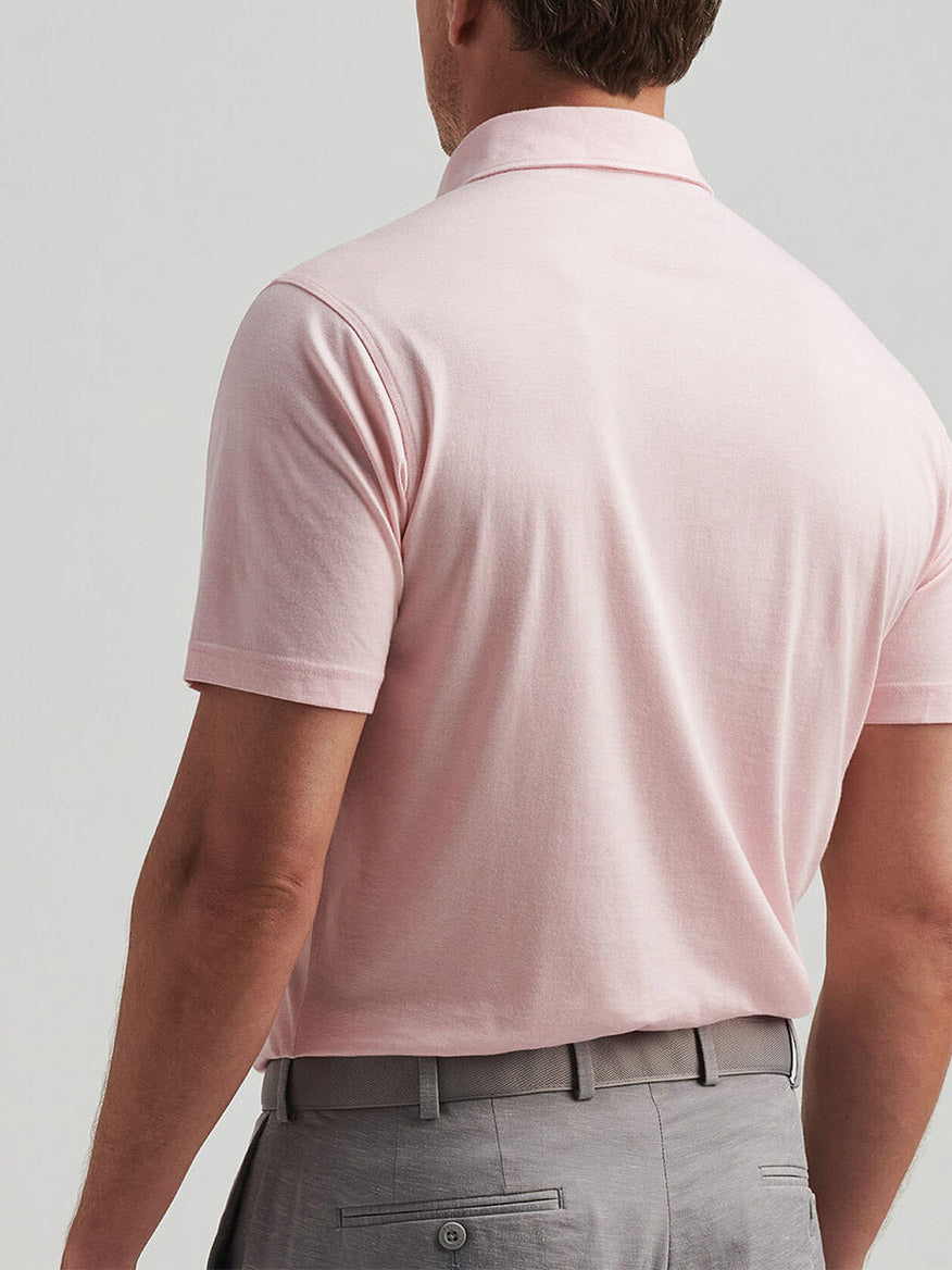 A man faces away, wearing a Peter Millar Excursionist Flex Polo in Pink Crepe, featuring a heathered design and tucked into light gray pants with belt loops.