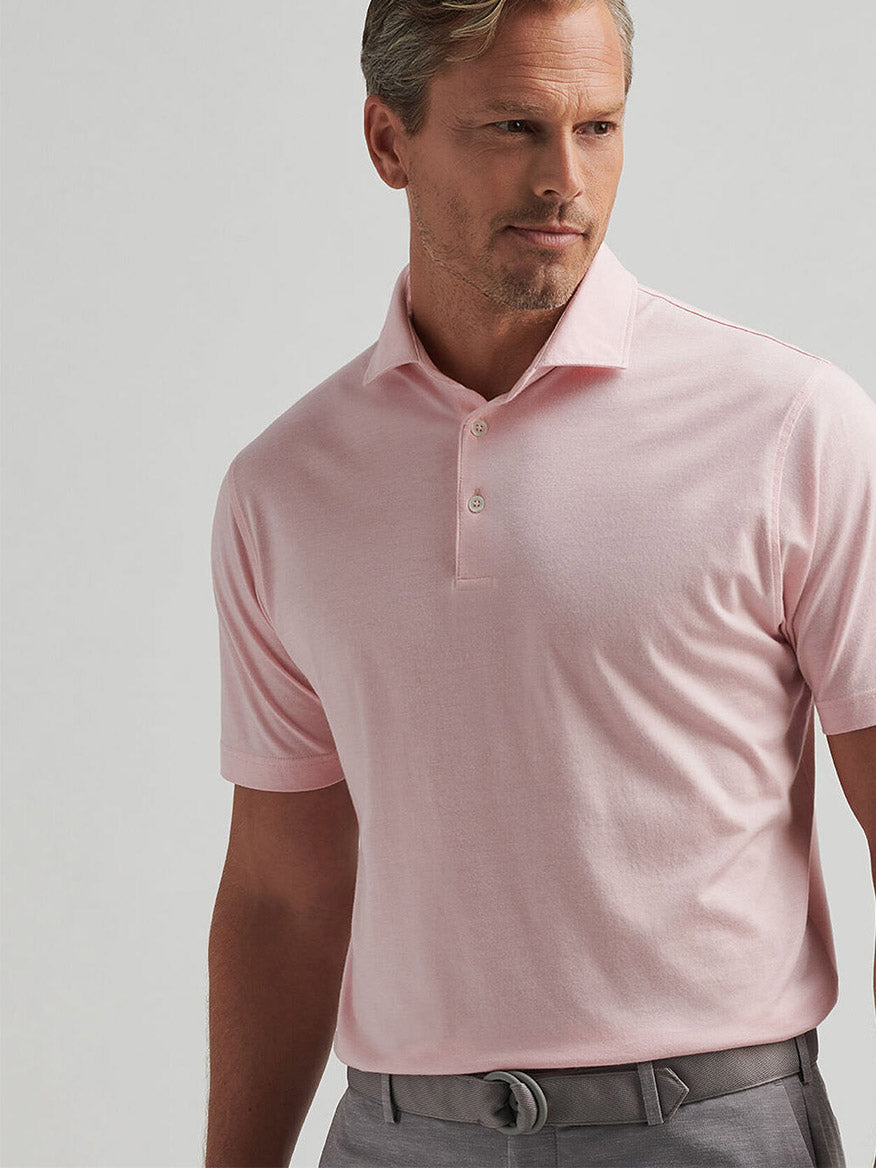 A man in a Pink Crepe Peter Millar Excursionist Flex Polo and gray pants stands facing slightly to the side against a plain background.