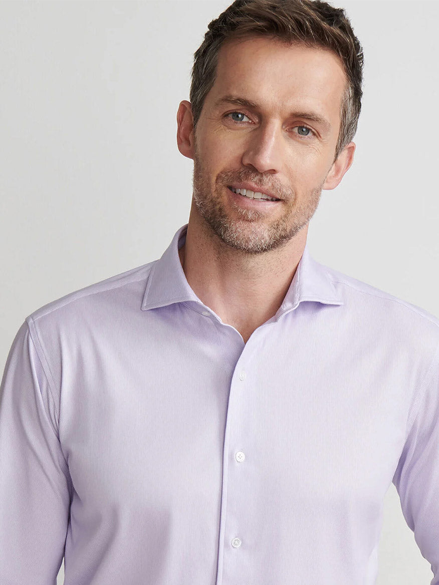 Man wearing a Peter Millar Excursionist Flex Micro Print Shirt in Wisteria, featuring cotton-stretch fabric, wrinkle resistance, and a tailored fit, stands against a plain light background and smiles at the camera.