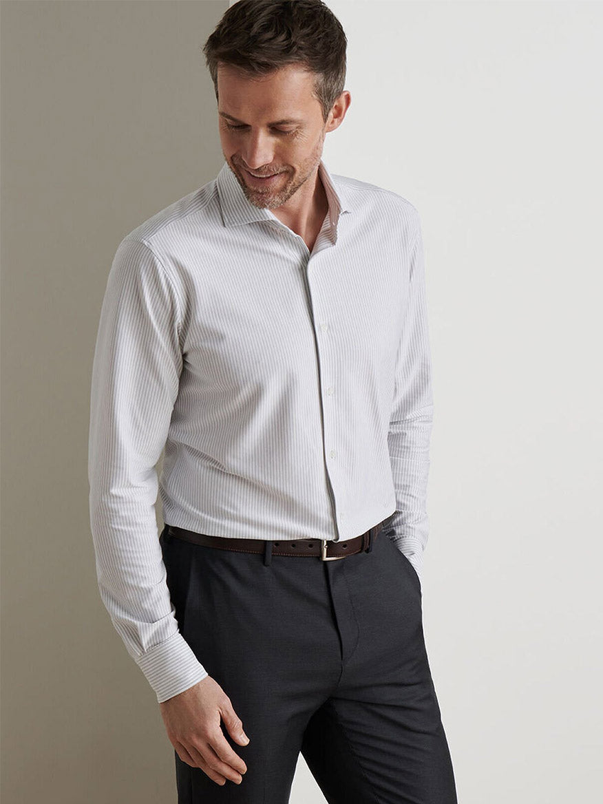 A man wearing a Peter Millar Excursionist Flex Stripe Sport Shirt in British Grey and dark trousers stands with one hand in his pocket, looking down and smiling.