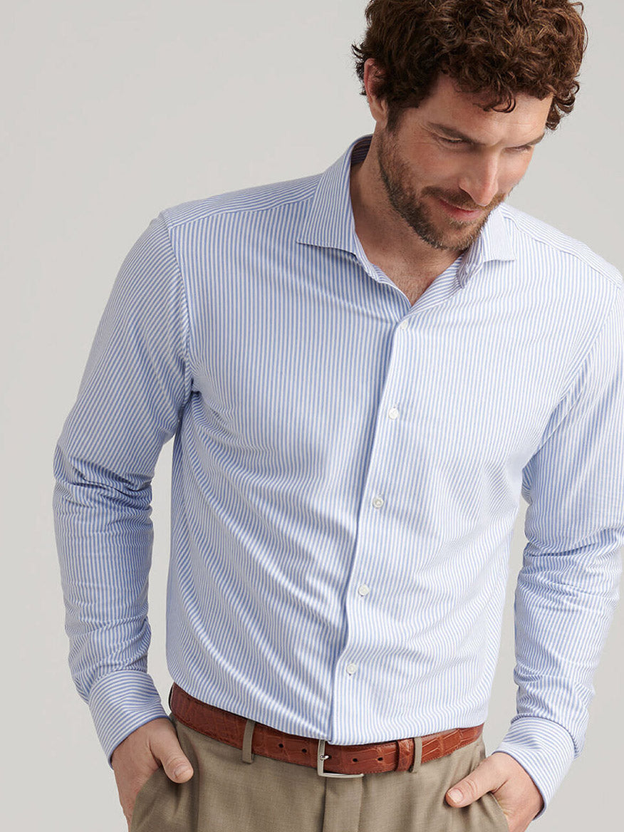 Man wearing the Peter Millar Excursionist Flex Stripe Sport Shirt in Rain Cloud, tucked into khaki pants with a brown belt, looking down and smiling slightly.