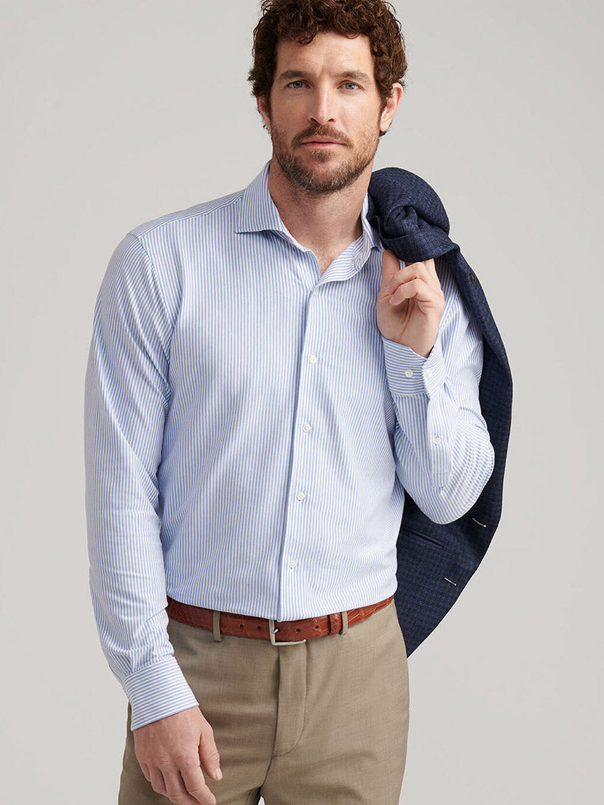 Man wearing a Peter Millar Excursionist Flex Stripe Sport Shirt in Rain Cloud with khaki pants, holding a navy blazer over his shoulder, standing against a plain background.