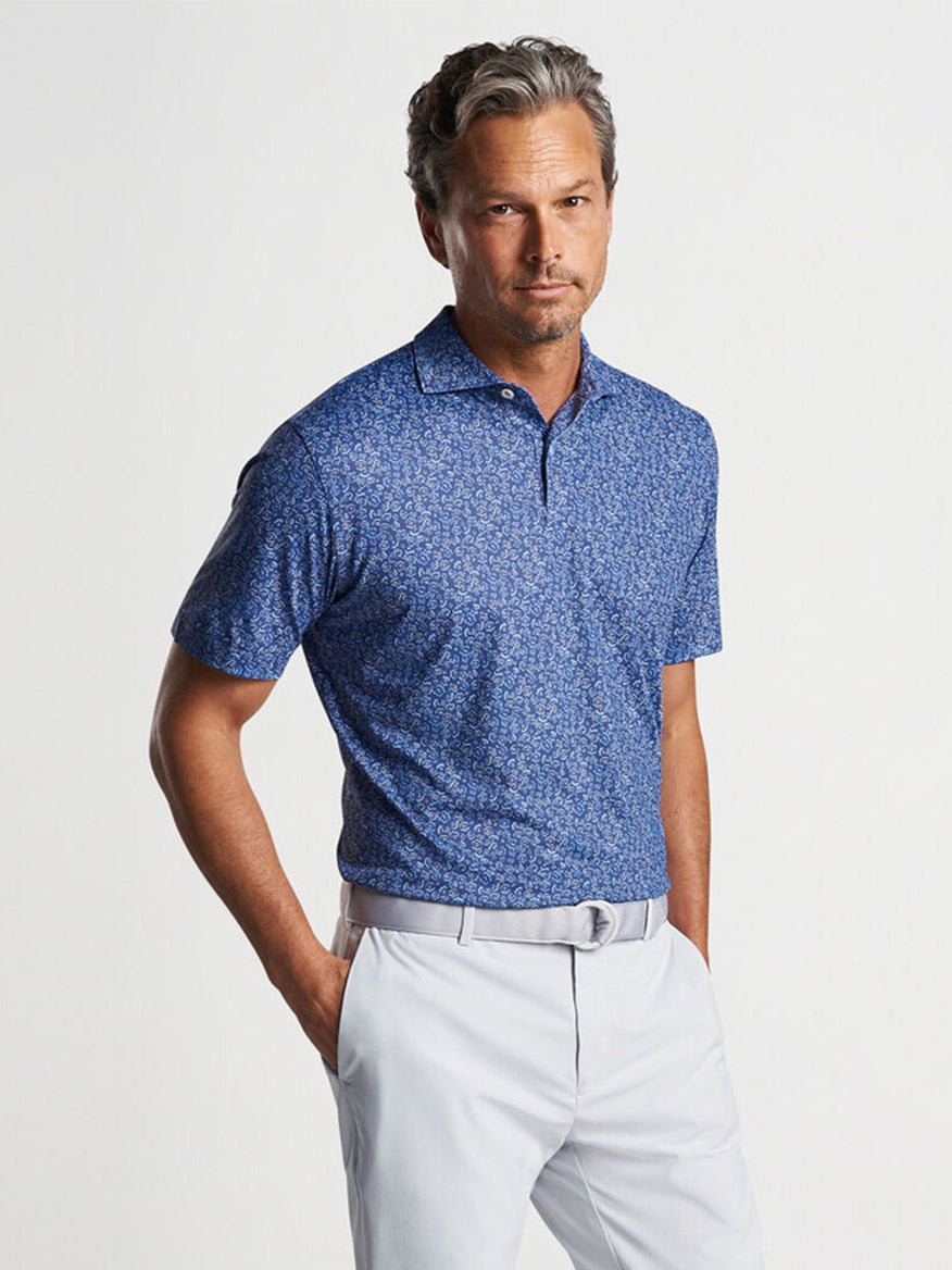 A man posing in a Peter Millar Fields of Carlsbad Performance Jersey Polo in Blue Pearl with UPF 50+ sun protection and light gray tailored fit pants with a belt.