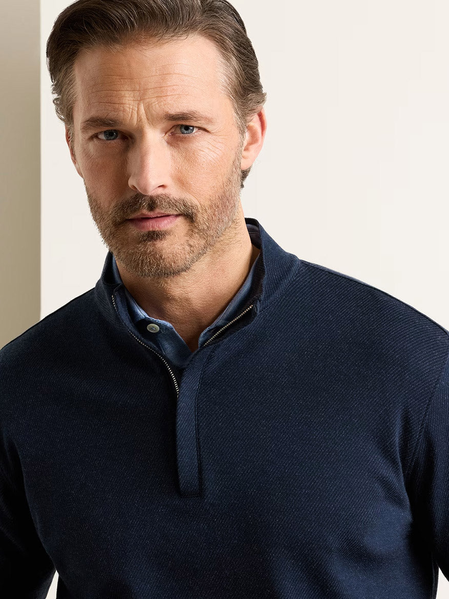 Man with light brown hair and beard wearing the Peter Millar Grant Knit Quarter-Zip in Navy, crafted from a soft cotton-cashmere blend, over a blue collared shirt, standing against an off-white background.