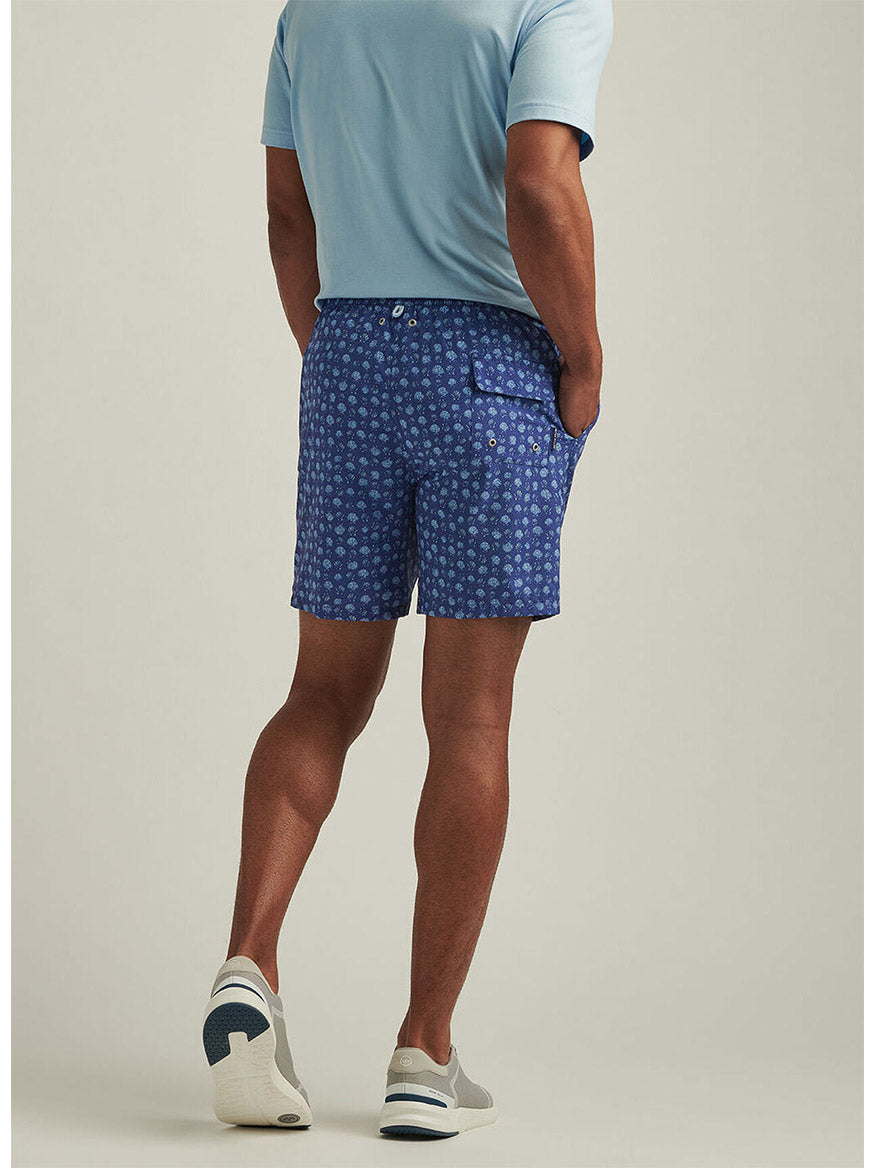 Sporting a Peter Millar Hatteras Harvest Swim Trunk in Ocean Blue, a person in a light blue shirt and sneakers stands with hands in pockets, facing away.
