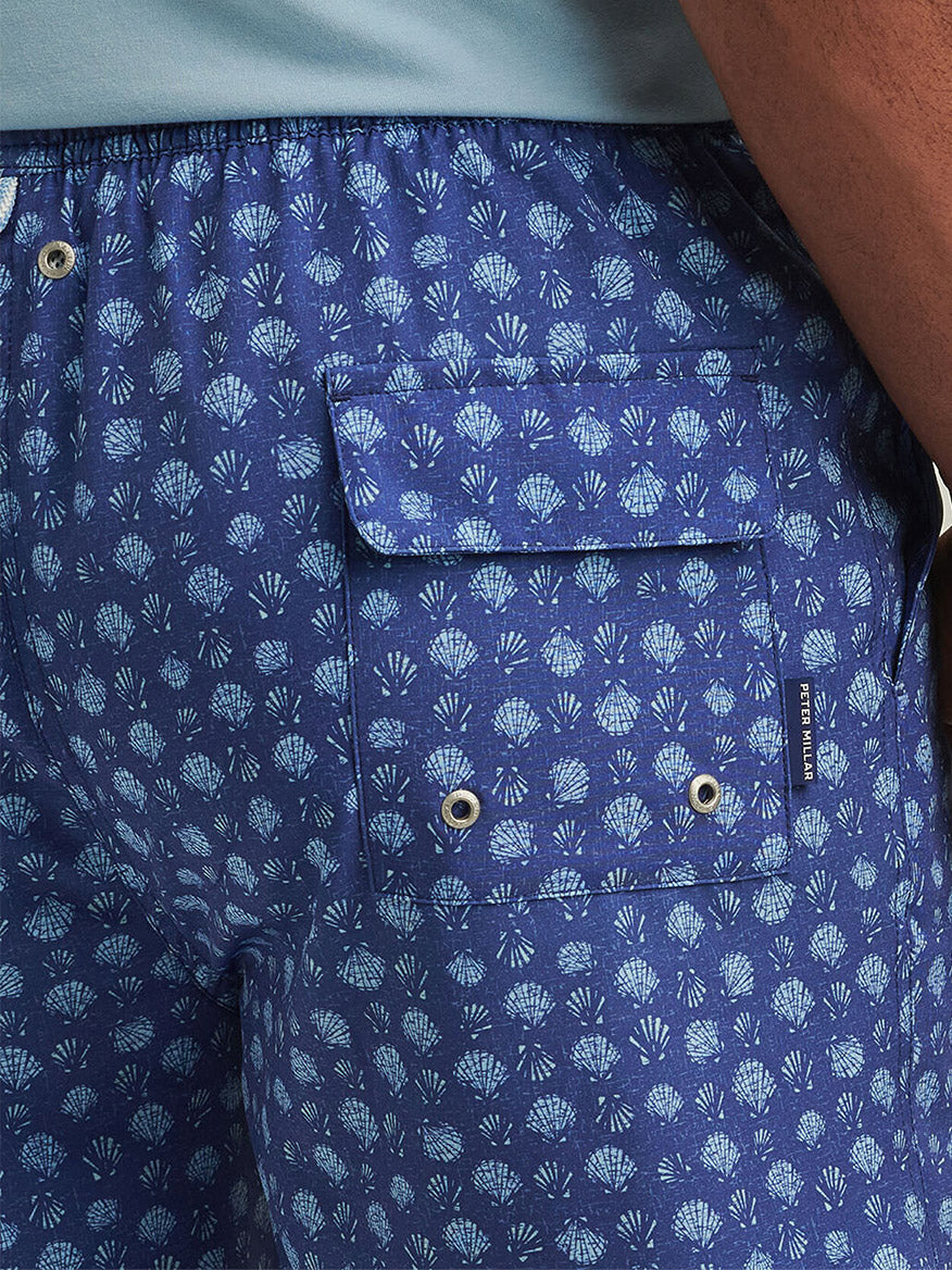Close-up of a person wearing the Peter Millar Hatteras Harvest Swim Trunk in Ocean Blue, featuring a vibrant seashell print, side buttoned pocket, and four-way stretch for comfort and mobility.