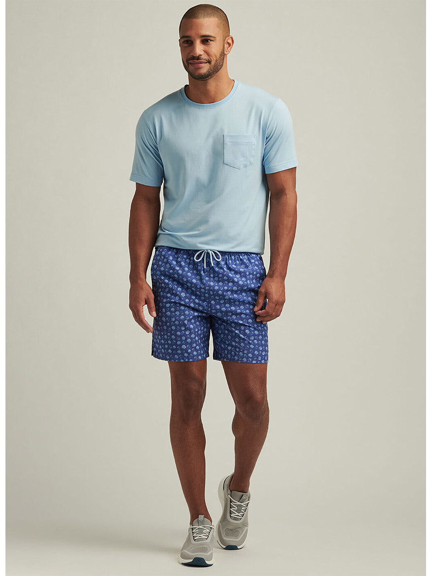 A man in a light blue T-shirt with a pocket, wearing Ocean Blue Peter Millar Hatteras Harvest Swim Trunks with seashell prints, stands against a neutral background. He completes the look with gray sneakers.