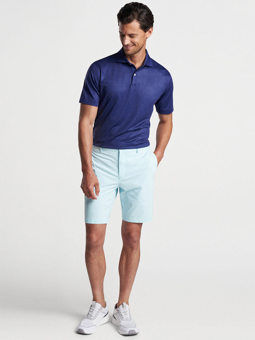 Man posing in a navy Peter Millar Instrumental Nouveau Performance Jersey Polo shirt with UPF 50+ sun protection, light blue shorts, and white sneakers.