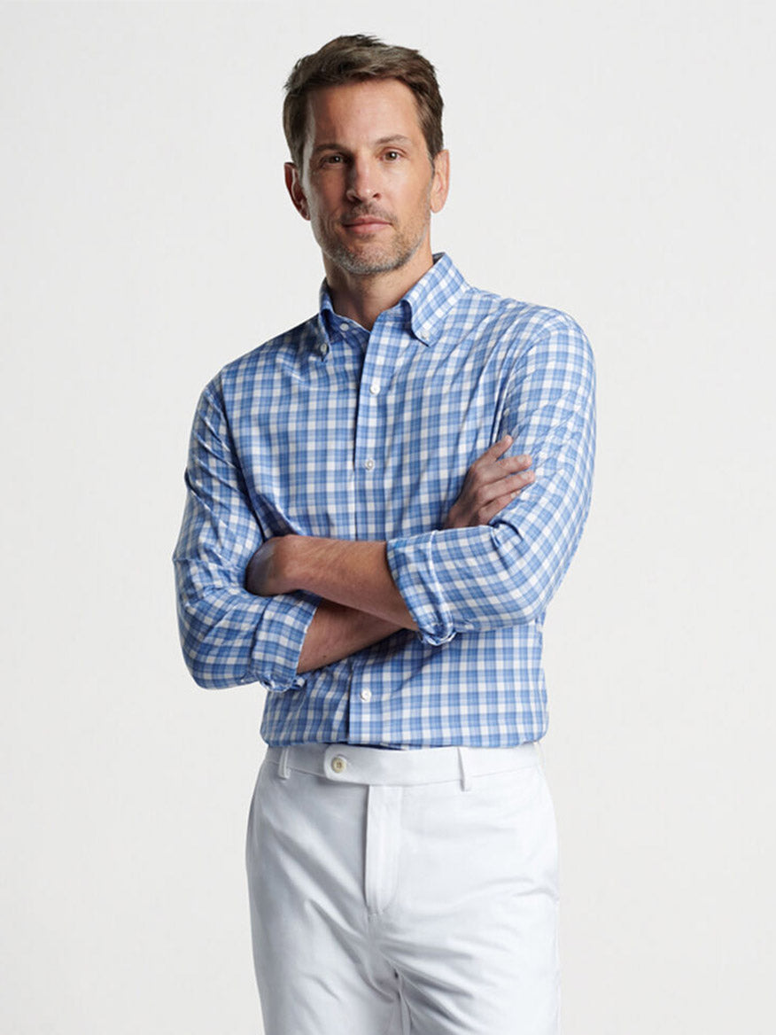 Man standing confidently with arms crossed, wearing a **Peter Millar Joplin Performance Poplin Sport Shirt in Regatta Blue** and white pants with UPF 50+ sun protection.