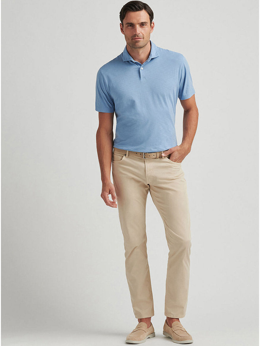 A man stands against a plain background wearing the Peter Millar Journeyman Short Sleeve Polo in Angelite, paired with beige pants, a brown belt, and beige loafers for a polished yet relaxed look.