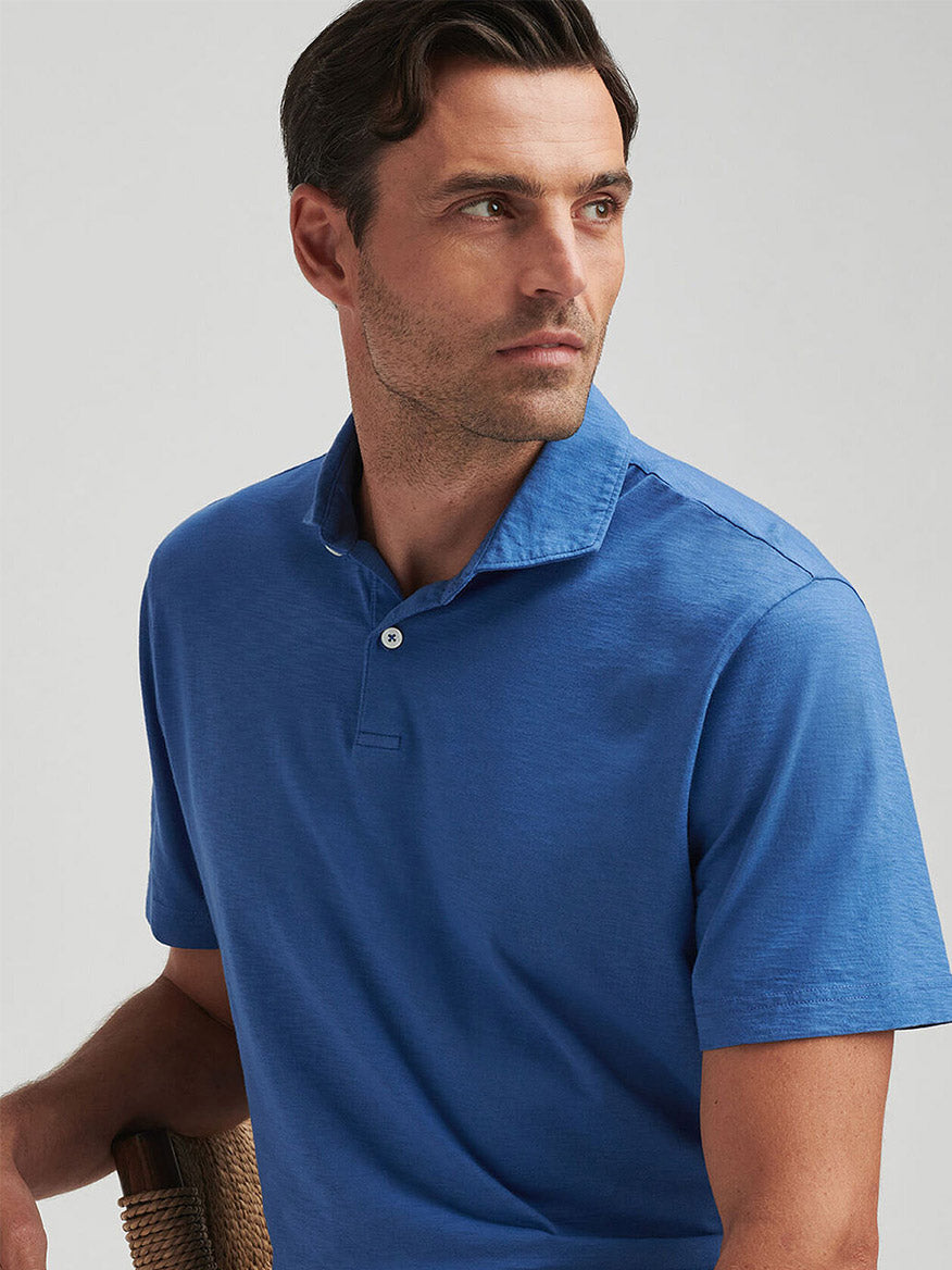 A man wearing the Peter Millar Journeyman Short Sleeve Polo in Ocean Blue with an Italian cutaway collar sits on a chair, looking left against a plain light background.