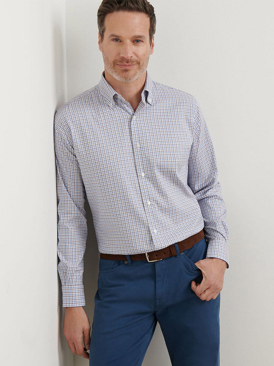 Man wearing the Peter Millar Julian Crown Lite Cotton-Stretch Shirt in Evening Tide, tucked into blue pants, leans against a white wall with one hand in his pocket. The easy-care fabric keeps him looking sharp all day.