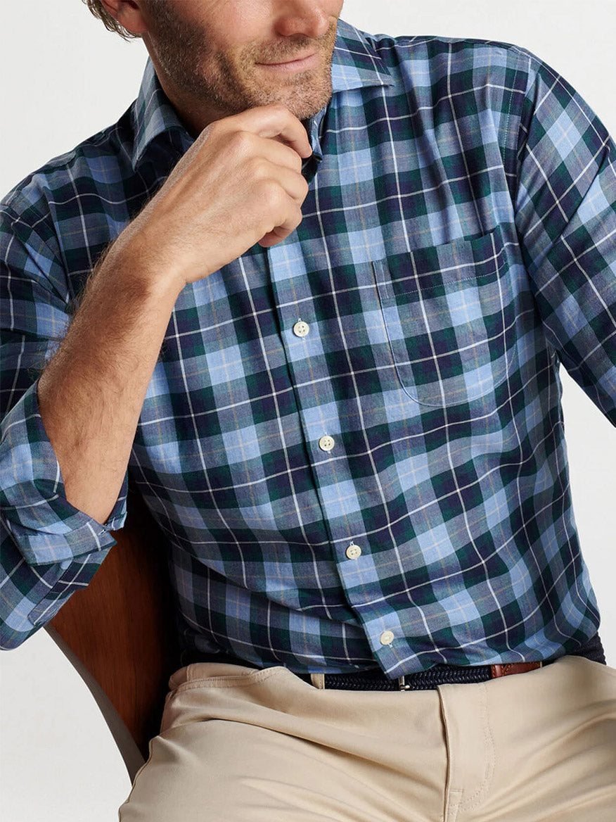 A person wearing a Peter Millar Langley Summer Soft Cotton Sport Shirt in Cottage Blue and beige cotton twill pants is seated with a hand at their chin.