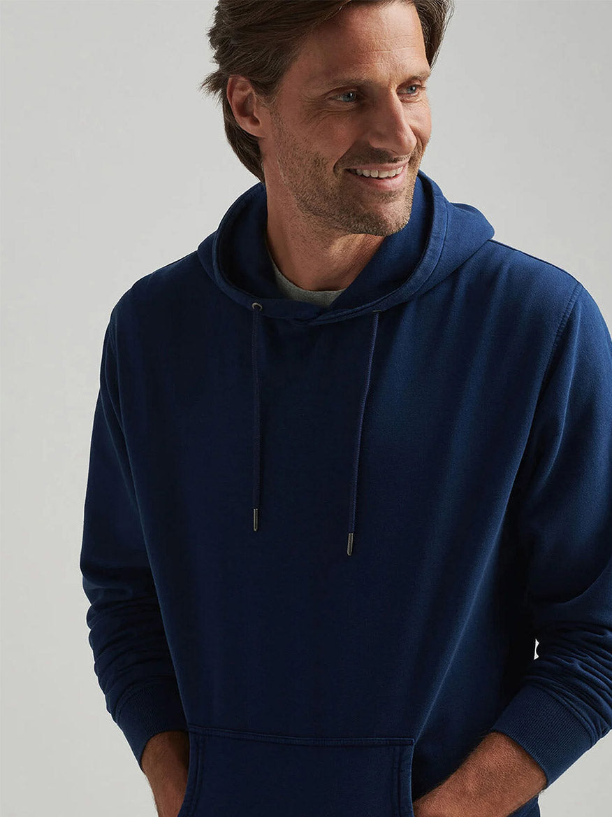 A man wears a Peter Millar Lava Wash Hoodie in Navy, smiling with his hands in the pockets and looking to the side against a plain background.