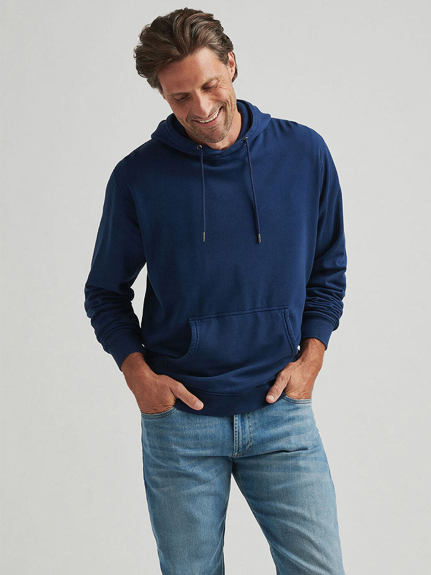 A man stands against a plain background, wearing the Peter Millar Lava Wash Hoodie in Navy with his hands in his pockets, paired with light blue jeans. He looks down and smiles.