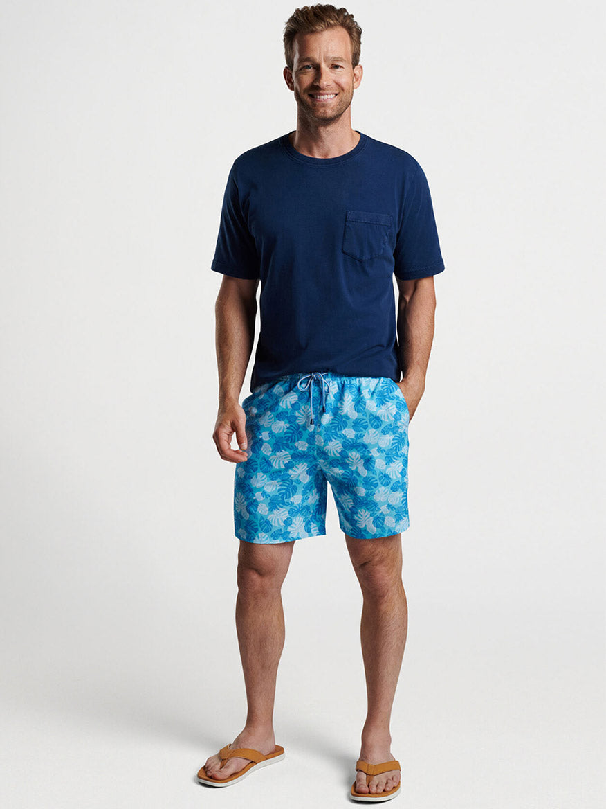 Man smiling in blue quick-drying fabric t-shirt and Peter Millar Linework Monstera Swim Trunk in Seasalt with flip-flops.