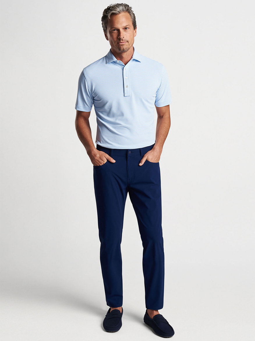 A man in a Peter Millar Mood Performance Mesh Polo in Blue Frost made of wicking mesh fabric and navy blue trousers standing against a plain background.