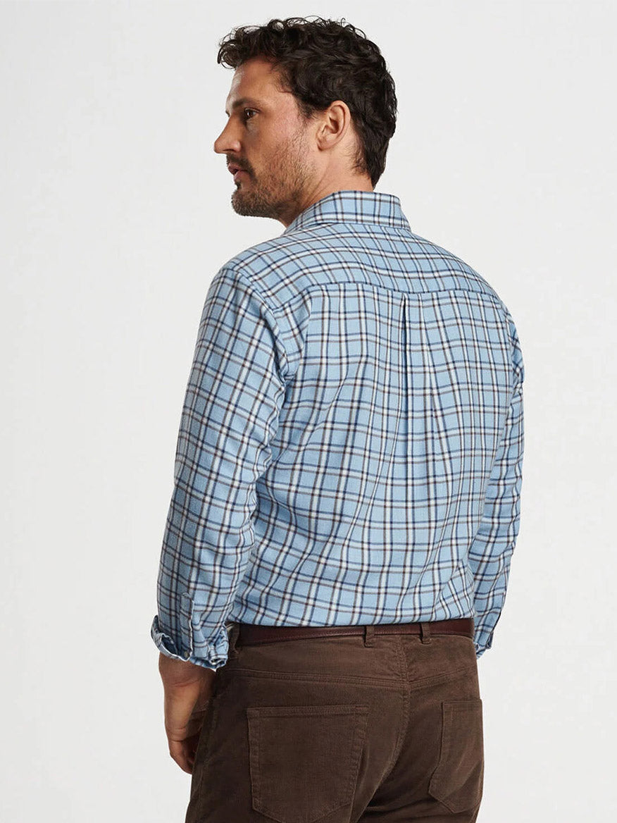 A man wearing the Peter Millar Nelson Flannelite Cotton-Stretch Sport Shirt in Cottage Blue paired with brown pants stands, facing slightly away from the camera. This shirt features a lightweight design with a touch of stretch, offering both style and practicality.