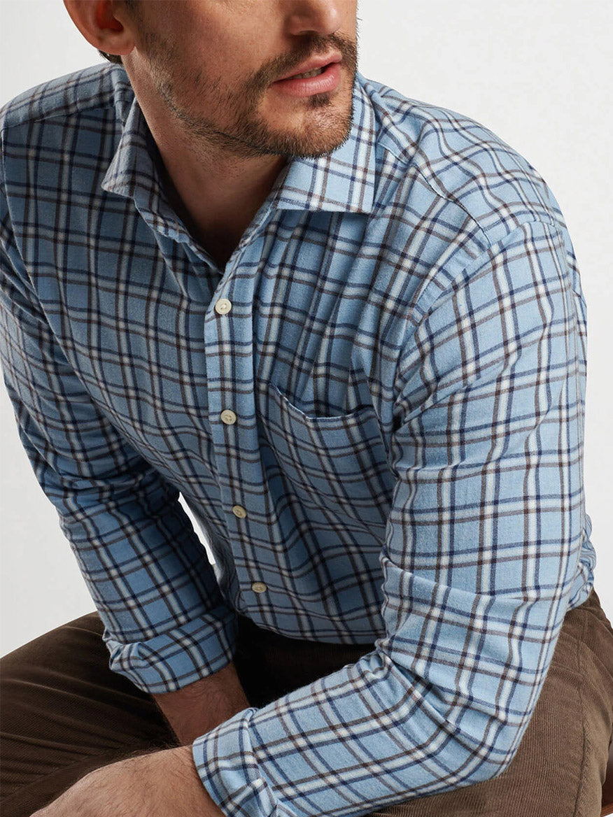 A man wearing a Peter Millar Nelson Flannelite Cotton-Stretch Sport Shirt in Cottage Blue and brown pants, leaning slightly forward with his arms resting on his knees.