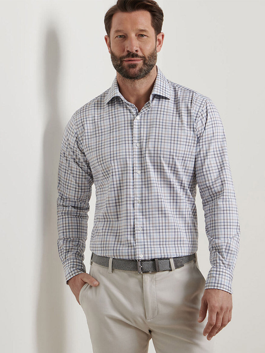 A bearded man wears a Peter Millar North Coast Crown Lite Cotton-Stretch Sport Shirt in Blue Haze, tucked into beige pants with a woven belt, standing against a plain white background.