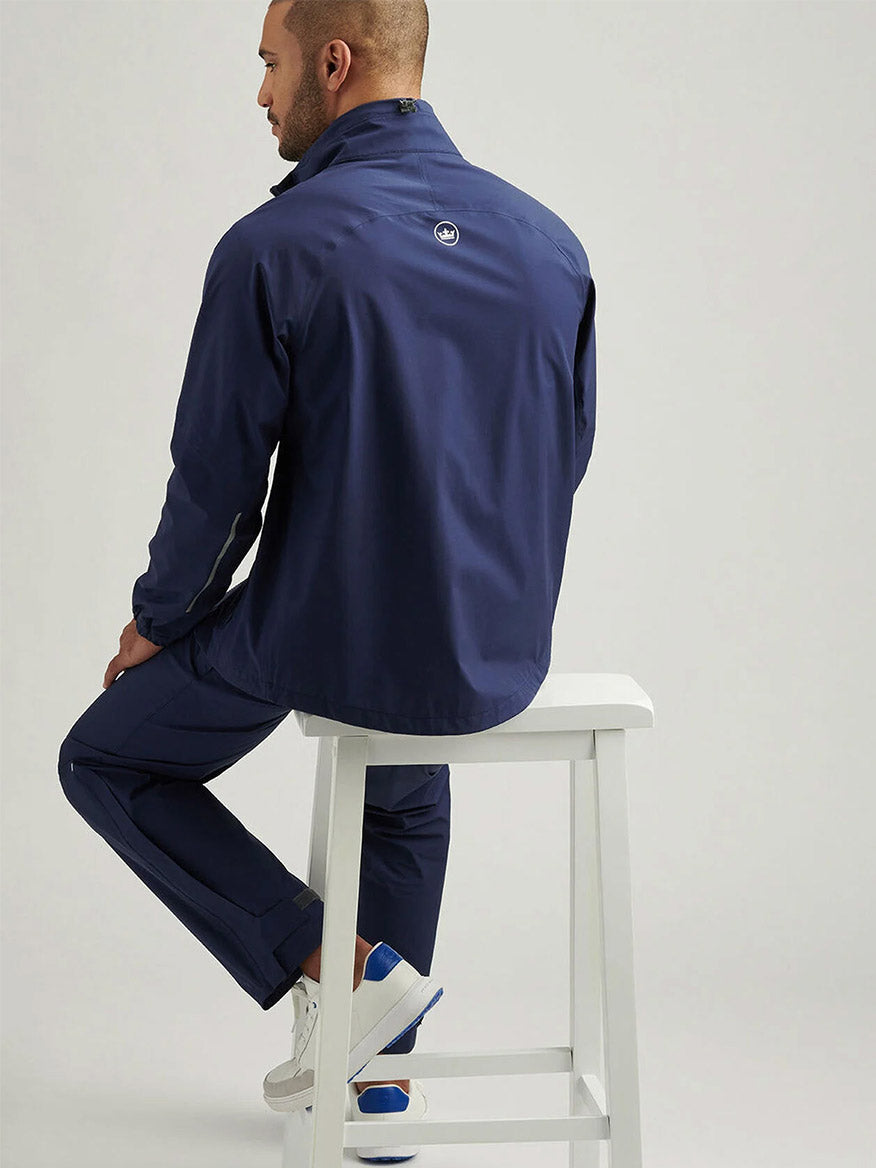 A man wearing the Peter Millar Rain Walker Pant in Navy sits on a white stool, facing away from the camera, against a plain light-colored background.