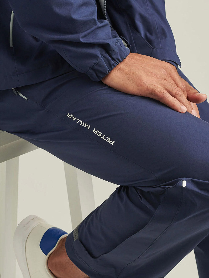 A person wearing the Peter Millar Rain Walker Pant in Navy, made from breathable fabric with "PETER MILLAR" branding, sits on a white stool with hands on their knee; white slip-on shoes featuring blue details are also visible.