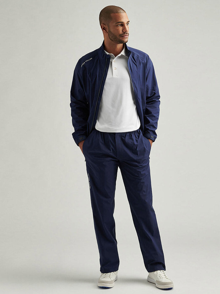 A man stands against a plain background wearing Peter Millar Rain Walker Pants in Navy, paired with a white polo shirt and white sneakers, with his hands in his pockets.
