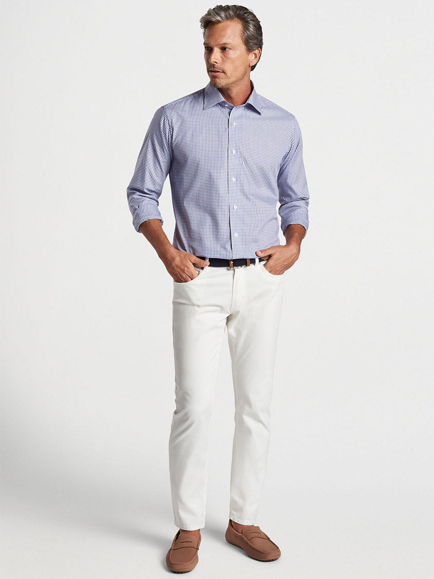 A man stands wearing a Peter Millar Renato Cotton Sport Shirt in Atlantic Blue, white cotton twill pants, and brown loafers, his hands resting on his hips in a studio setting.