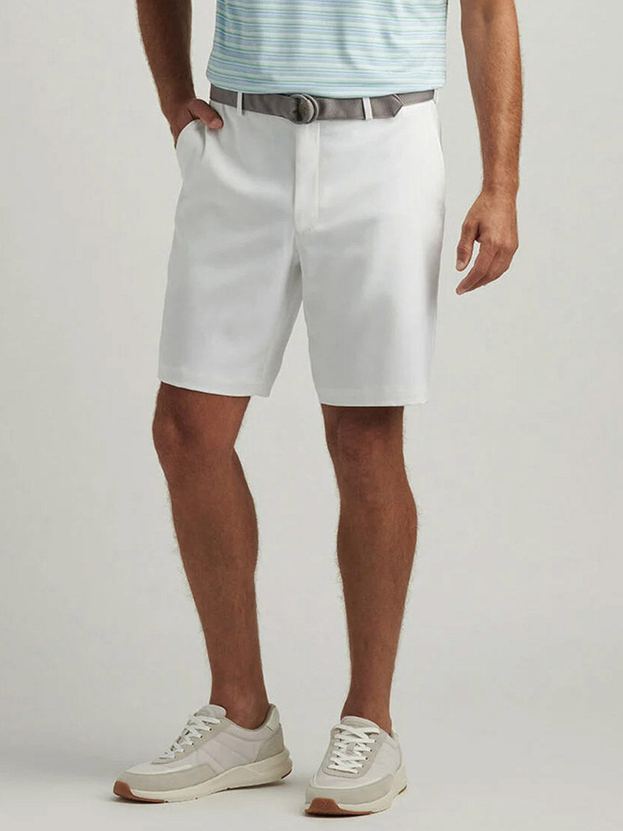 A man wearing a light blue striped polo, Peter Millar Salem Performance Short in White with a gray belt, and white sneakers stands with one hand in his pocket.