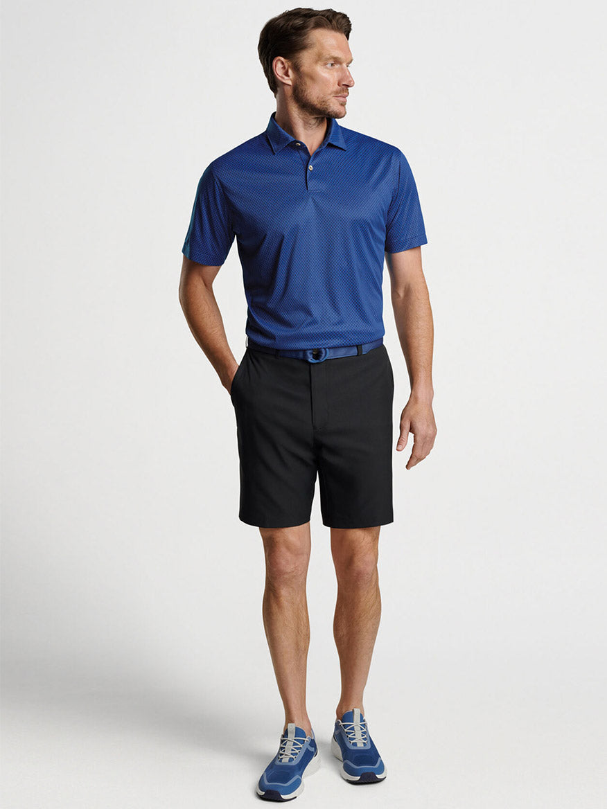 Man wearing a blue golf shirt made of quick-dry performance fabric and Peter Millar Salem Performance Short in Black with matching blue golf shoes.