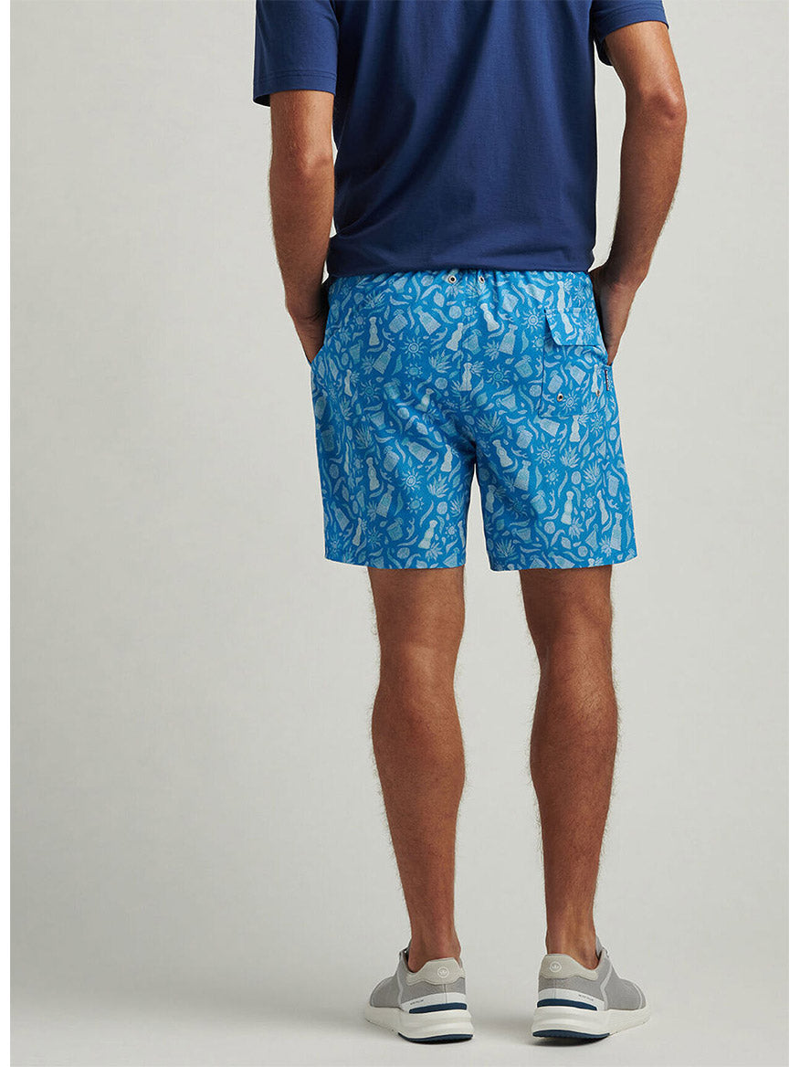 A man stands with his back to the camera, sporting a navy shirt and Peter Millar Saltwater Spirits Swim Trunk in Blue Macaw. His ensemble, completed by gray sneakers, showcases unique style flair and comfort.