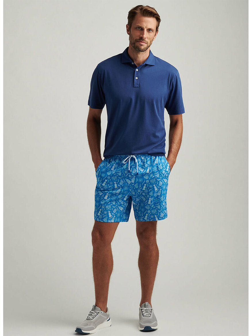A man in a blue polo with the Peter Millar Saltwater Spirits Swim Trunk in Blue Macaw—featuring an exclusive hand-designed print—stands with hands in his pockets, pairing the look with gray sneakers.
