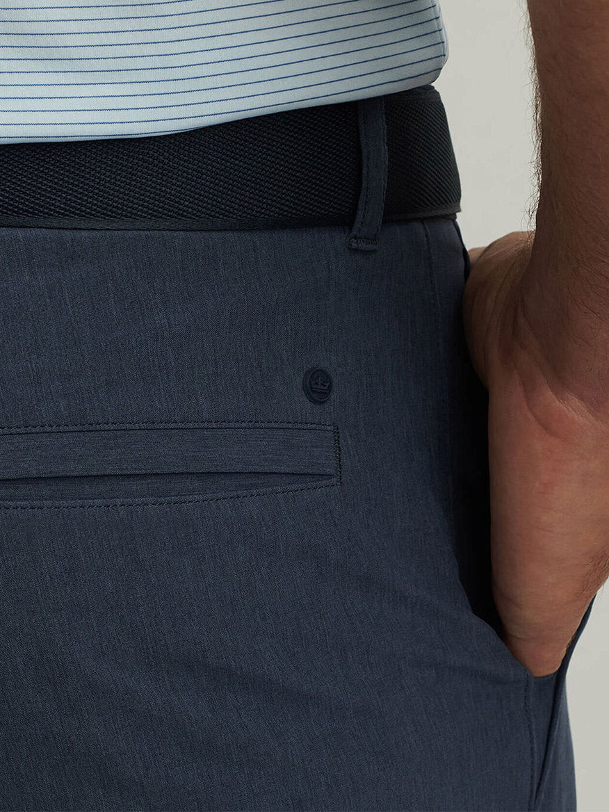 Wearing the Peter Millar Shackleford Performance Hybrid Short in Navy with four-way stretch and a black belt, this person pairs them with a light blue striped shirt, right hand tucked casually in their pocket.