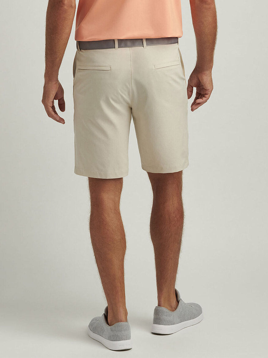 A man wearing a peach polo shirt, Peter Millar Shackleford Performance Hybrid Short in Sand, and gray slip-on shoes stands facing away from the camera against a plain background.