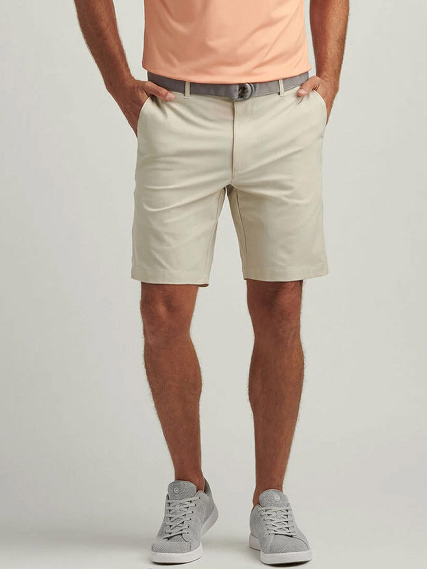 A person wearing a peach shirt, Peter Millar Shackleford Performance Hybrid Short in Sand, a gray belt, and gray sneakers stands with hands in pockets against a light background.