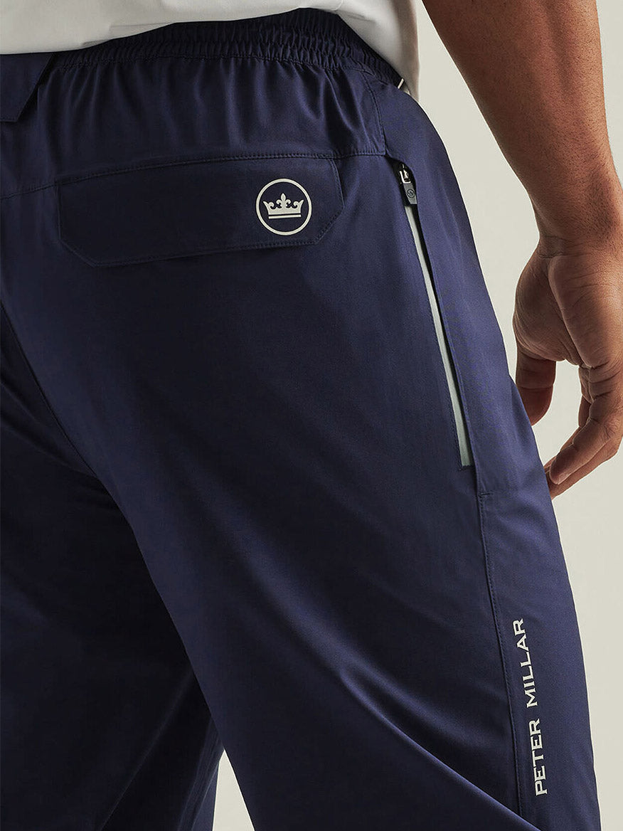 A person wearing navy blue Peter Millar Shield Pull-On Rain Pants, featuring technical performance fabric, a zippered pocket, and a visible crown logo near the waistband.